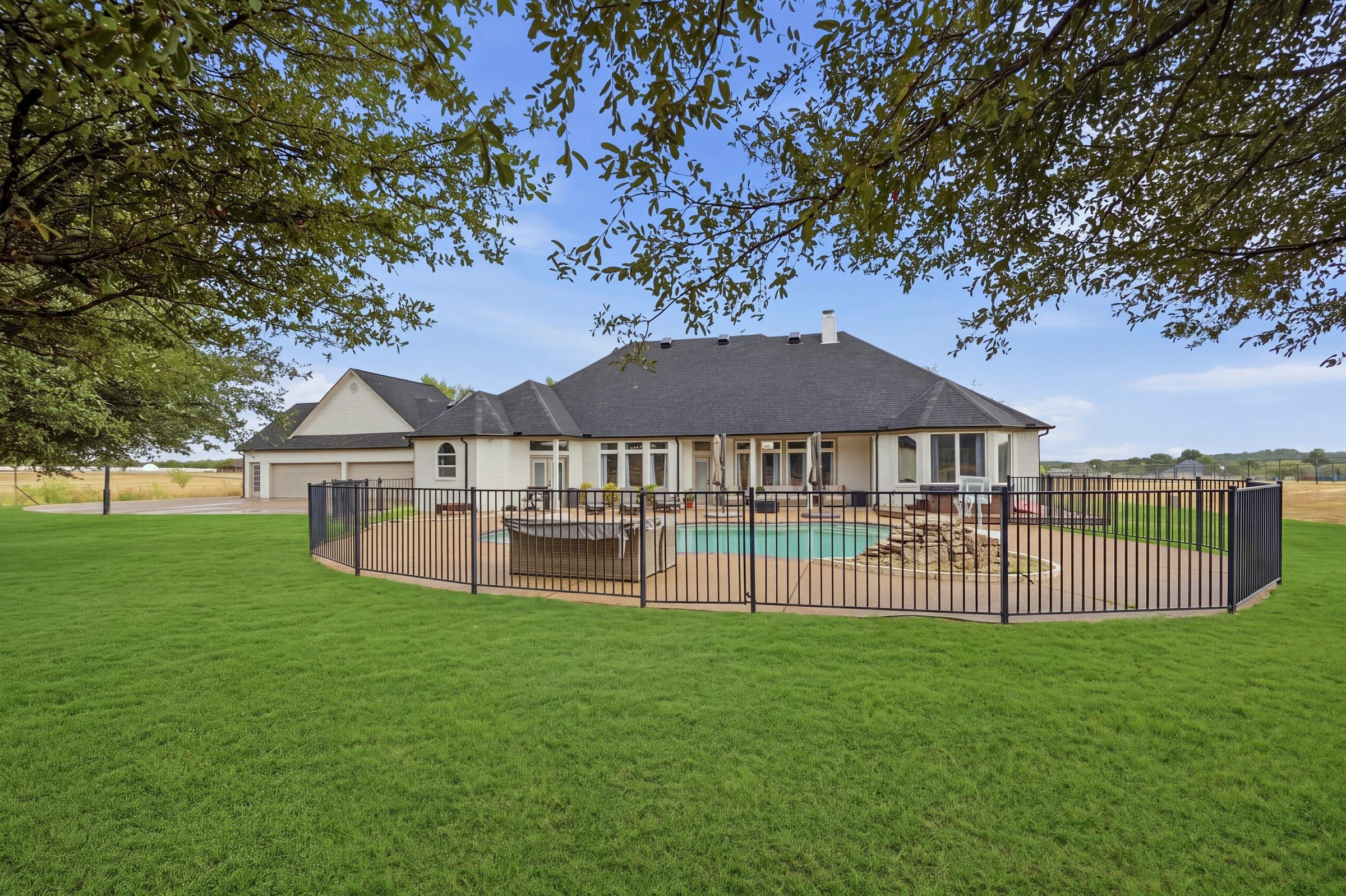 Fenced pool and back of the home seen from the yard under an oak tree